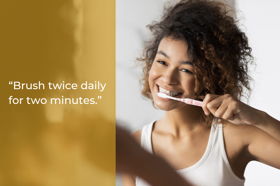 A woman smiles while brushing her teeth in front of a mirror; text on the left reads, “Brush twice daily for two minutes—oral hygiene starts with simple home tips.”.